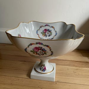以下が含まれることがあります： 白い磁器製のセンターピースボウル。縁はスカラップで、金色のトリムが付いています。ボウルにはピンク、青、黄色の花柄のデザインが施されています。正方形の台座に置かれており、これも白で金色のアクセントが付いています。装飾品です。