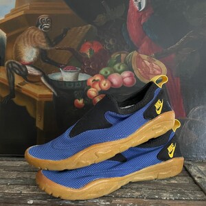 May include: Blue and black slip-on shoes with a tan sole. The shoes have a mesh-like texture on the blue portion and a yellow Nike logo. The shoes are stacked on top of each other.