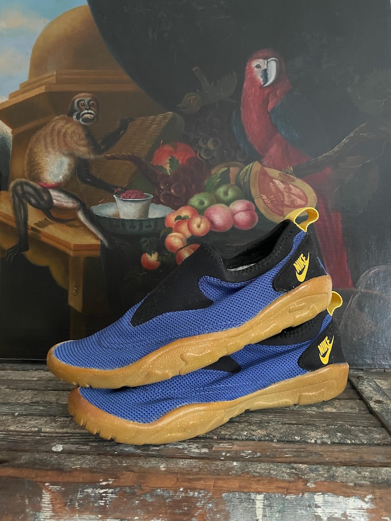 May include: Blue and black slip-on shoes with a tan sole. The shoes have a mesh-like texture on the blue portion and a yellow Nike logo. The shoes are stacked on top of each other.