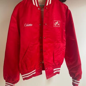 May include: Red satin baseball jacket with white and red striped ribbing. The jacket has a button closure and features the text "Colette" embroidered on the left side and a logo with the text "Like" on the right side.