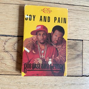 May include: A retro cassette tape with the title "JOY AND PAIN" in black text on a yellow background. The cover features two men, one wearing a red Ferrari cap. The artist's name, "ROB BASE & D.J. EZ ROCK," is also displayed.
