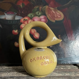 Könnte beinhalten: Ein gelber Keramik Krug mit einem einzigartigen Griff in Form eines Vogelhalses. Der Krug trägt die Aufschrift "OKARCHE OKLA." auf der Seite.