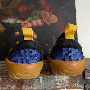 May include: A pair of children's shoes with a black upper, blue mesh detailing, and a tan sole. The shoes have a yellow pull tab on the back. The shoes are designed for comfort and support.