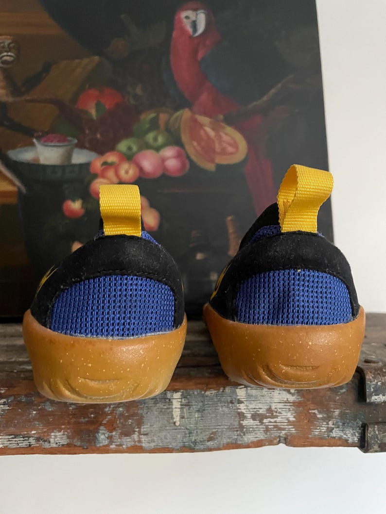 May include: A pair of children's shoes with a black upper, blue mesh detailing, and a tan sole. The shoes have a yellow pull tab on the back. The shoes are designed for comfort and support.