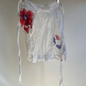 May include: White linen apron with red and blue floral print. The apron has two long ties and a small pocket.