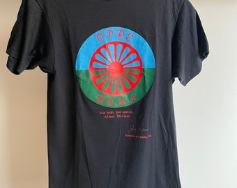 May include: Black t-shirt with a circular graphic featuring a red sun with green and blue rings around it. The text "OPRE ROMA" is printed in red letters. The text "hut bakt, but sastipe Achen 'Devlesa'" is printed in black letters below the graphic. The text "ROMAMA OF MASS. INC." is printed in black letters below the text.
