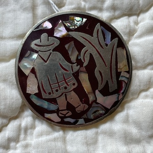 May include: A round, silver-toned pendant with a burgundy background. The pendant features a stylized image of a person in a hat and a long coat, standing next to a plant. The image is made of iridescent mother-of-pearl pieces.