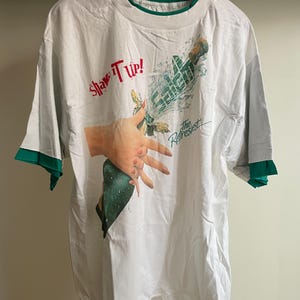 May include: White t-shirt with green trim featuring the text "Shake It Up!" in red and "The Refreshiest" in blue. The graphic shows a hand holding a bottle with water splashing out.
