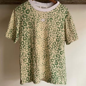 May include: A short-sleeved t-shirt featuring a green leopard print pattern on a cream background. The garment has a solid white crew neckline and a small white embroidered logo on the chest. The interior neck label displays the text Wales Bonner.