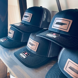 May include: A shelf display of black and grey trucker hats with a brown leather patch featuring the outline of the state of Tennessee. The hats have a mesh back and a white stitching detail.