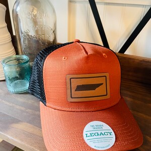 May include: A brown and black trucker hat with a brown leather patch featuring the outline of the state of Tennessee. The hat has a black mesh back and a white sticker with the text "The Roadie Trucker Legacy" and the text "Premium, Vintage-Inspired Caps for a Relaxed Lifestyle #LiveYourLegacy".