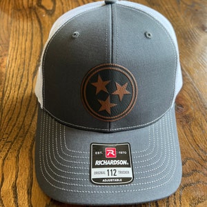 May include: Gray and white trucker hat with a brown leather patch featuring a three-pointed star design. The patch has the text "EST. 1970 RICHARDSON. ORIGINAL 112 TRUCKER ADJUSTABLE" on it.