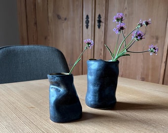 Könnte beinhalten: Zwei dunkelblaue Keramikvasen mit einer strukturierten, unebenen Oberfläche. Die höhere Vase enthält einen Strauß lila Blumen.