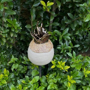 Könnte beinhalten: Ein dekorativer Gartenstecker mit einer cremefarbenen Kugelbasis, einem gemusterten Band und einer bronzefarbenen floralen Spitze. Der Stecker befindet sich vor einem Hintergrund aus grünem Laub und schafft eine natürliche, äußere Ästhetik.