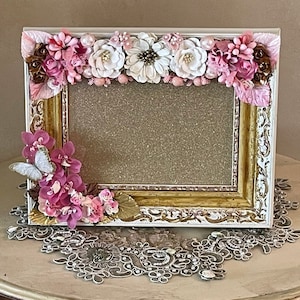 May include: A rectangular picture frame with a gold and white ornate design, adorned with pink and white paper flowers, leaves, and pearls. A butterfly rests on the lower left corner. The frame is displayed on a decorative table.