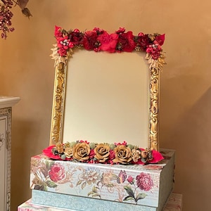 May include: A gold-framed mirror adorned with red and gold floral arrangements, resting atop three decorative floral-patterned boxes. The boxes are stacked, showcasing a variety of floral designs and colors, including pink, blue, and cream.