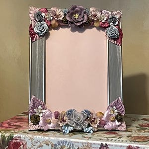 May include: A silver-framed picture frame with a pink interior, adorned with an array of fabric flowers in shades of purple, pink, and gray. The frame sits atop a decorative box with floral and butterfly illustrations.