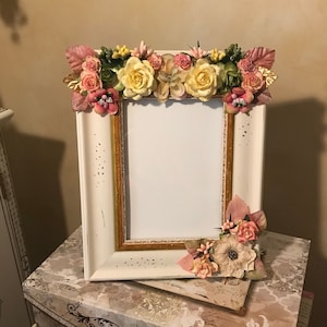 May include: A white rectangular picture frame with a gold trim, adorned with an array of artificial flowers in shades of pink, yellow, and green. The frame is placed on top of decorative boxes with floral and butterfly patterns.