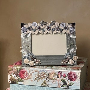 May include: A decorative silver picture frame adorned with floral embellishments in shades of blue, white, and gray. The frame sits atop a stack of floral-patterned boxes in pink, blue, and white, creating a vintage aesthetic.