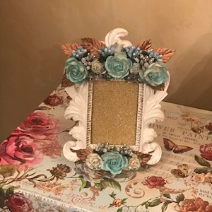 May include: A white, ornate picture frame with a gold glitter insert, adorned with teal roses and brown leaves. The frame sits atop a floral-patterned box with roses and butterflies.