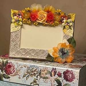 May include: A decorative photo frame with a floral arrangement of yellow, orange, and pink flowers. The frame has a textured, off-white surface and is adorned with leaves and additional floral accents. The frame sits atop floral-patterned boxes.