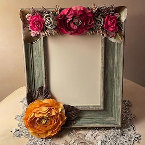 May include: A decorative picture frame with a distressed teal finish. The frame is adorned with fabric flowers in shades of pink, orange, and green. The frame is resting on a white lace doily.