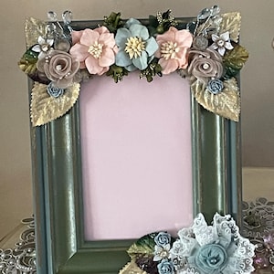 May include: A rectangular picture frame with a distressed green and blue finish. The frame is adorned with fabric flowers in shades of pink, blue, and cream, along with gold-colored leaves and crystal accents. The frame is designed to hold a photo.