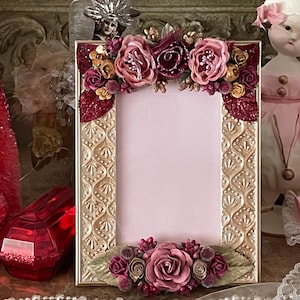 May include: A rectangular gold-colored picture frame with a pink interior. The frame is decorated with embossed patterns and adorned with fabric flowers in shades of pink, burgundy, and gold. The flowers are clustered at the top and bottom.