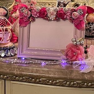 May include: A pink picture frame decorated with pink and red flowers, leaves, and silver accents. The frame is surrounded by decorative dolls and a string of purple lights. The frame is a decorative item.