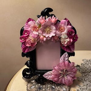 May include: A black ornate picture frame adorned with pink and fuchsia fabric flowers and embellishments. The frame is decorated with a floral arrangement across the top and a large flower on the side, creating a decorative display.