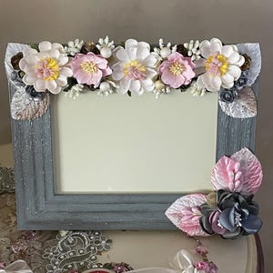 May include: A rectangular photo frame with a distressed gray finish, adorned with fabric flowers in white and pink. Decorative leaves and embellishments accent the corners. The frame is designed for displaying photographs.