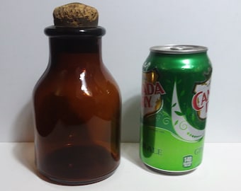 May include: A brown glass bottle with a cork stopper next to a green Canada Dry ginger ale can. The bottle has a rounded shape and a dark brown color. The can is green with white text and a leaf design.