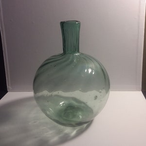 May include: A clear green glass bottle with a round body and a short neck. The bottle has a swirl pattern and is slightly textured.