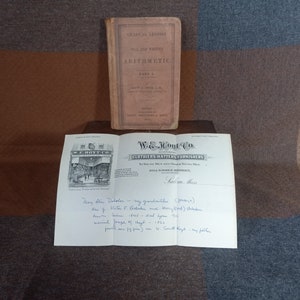May include: A vintage book titled "Gradual Lessons of Oral and Written Arithmetic, Part I" by John H. Walsh. The book is closed and sitting on a wooden surface. A folded piece of paper with a business card for W.E. Hoyt Co. Clothiers, Hatters, Furnishers, Salem, Mass. is in front of the book. The business card has a small illustration of the store front. The paper also has handwritten text.