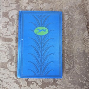 Peut inclure: Un livre vintage avec une couverture bleue ornée d'un motif décoratif et d'un ovale vert avec la silhouette d'un chien. La couverture du livre présente un motif floral détaillé dans une nuance de bleu plus foncée.