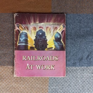 以下が含まれることがあります： ピンクの背景に白い文字で「Railroads At Work」と書かれた、ビンテージな本の表紙。表紙には、橋と街並みを背景に、中央に1つ、両側に1つずつ、合計3つの列車が描かれたイラストが描かれている。