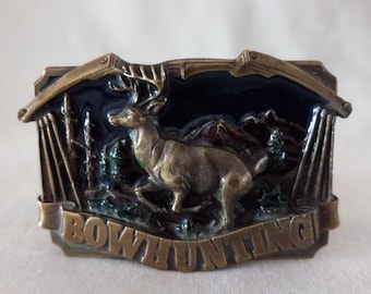 May include: A brass belt buckle featuring a deer and bow and arrow design. The buckle is enamelled in black, green, and red. The text "BOWHUNTING" is engraved on the buckle.