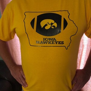May include: A yellow t-shirt featuring a black and gold design. The design includes a football shape with the outline of the state of Iowa, and the words "IOWA HAWKEYES" below the football.