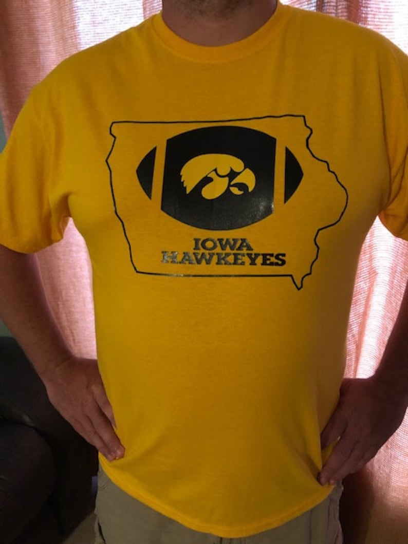 May include: A yellow t-shirt featuring a black and gold design. The design includes a football shape with the outline of the state of Iowa, and the words "IOWA HAWKEYES" below the football.