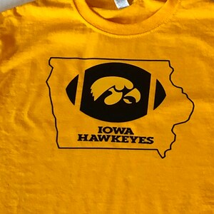 May include: A yellow t-shirt featuring a black and gold graphic of the state of Iowa with a football and the Iowa Hawkeyes logo. The text "IOWA HAWKEYES" is printed below the logo.