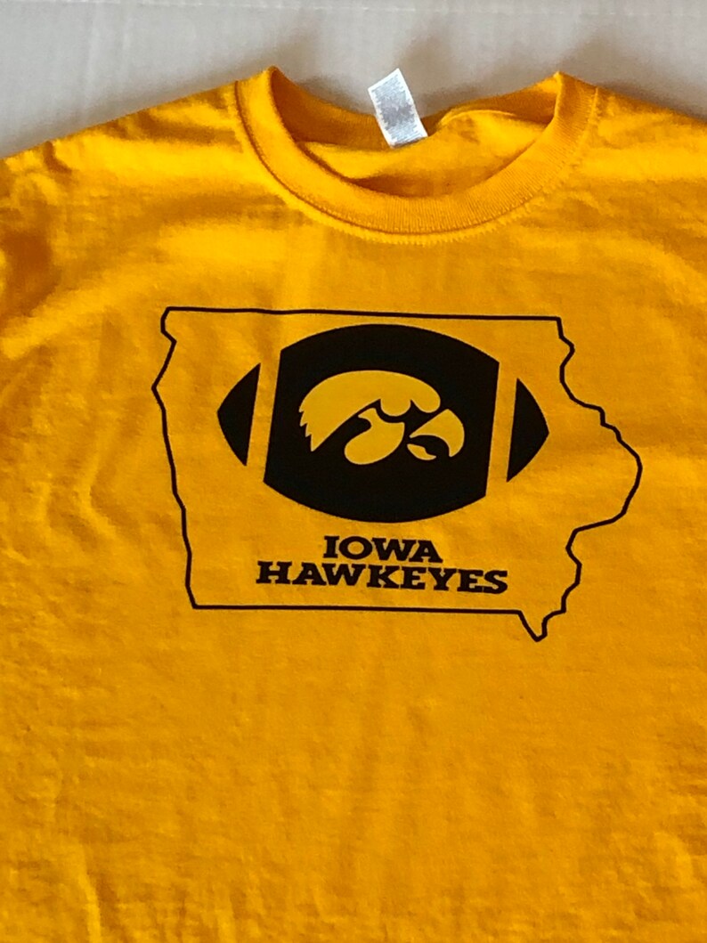 May include: A yellow t-shirt featuring a black and gold graphic of the state of Iowa with a football and the Iowa Hawkeyes logo. The text "IOWA HAWKEYES" is printed below the logo.