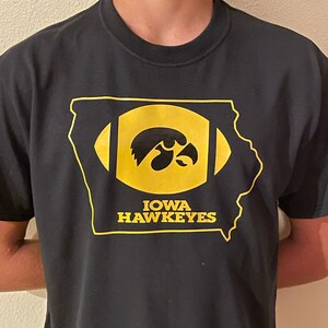 May include: Black t-shirt featuring a gold outline of the state of Iowa with a football design. Inside the football is the Iowa Hawkeyes logo and the words "IOWA HAWKEYES" in gold.