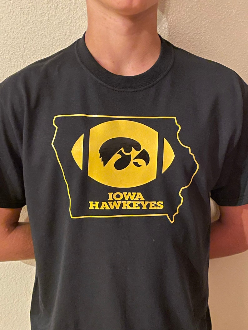 May include: Black t-shirt featuring a gold outline of the state of Iowa with a football design. Inside the football is the Iowa Hawkeyes logo and the words "IOWA HAWKEYES" in gold.