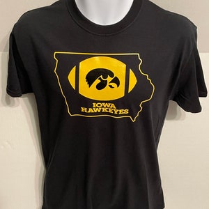 May include: Black t-shirt featuring a gold-colored design of the state of Iowa shaped like a football. Inside the football shape is the Iowa Hawkeyes logo, a hawk's head. The text "IOWA HAWKEYES" is below the logo.