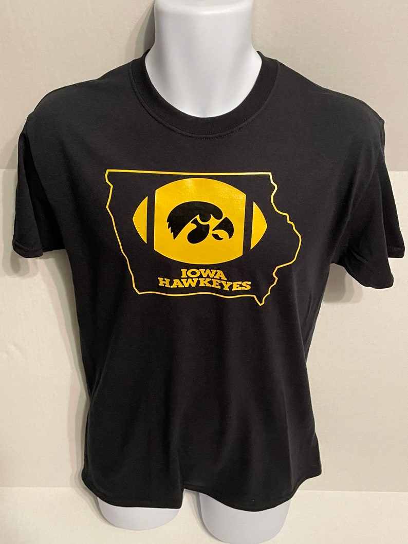 May include: Black t-shirt featuring a gold-colored design of the state of Iowa shaped like a football. Inside the football shape is the Iowa Hawkeyes logo, a hawk's head. The text "IOWA HAWKEYES" is below the logo.