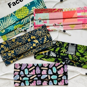 May include: Five different fabric face masks with various patterns. The masks are all folded and have white elastic ear loops. The patterns include pineapples, geometric shapes, flowers, sloths, and crystals.