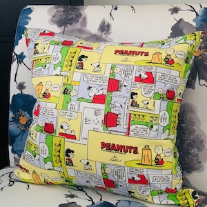 Puede incluir: Cojín decorativo cuadrado con un diseño de cómic colorido con personajes de Peanuts. El cojín tiene un fondo amarillo con paneles de texto e ilustraciones. El logotipo de Peanuts es visible. Decoración divertida.