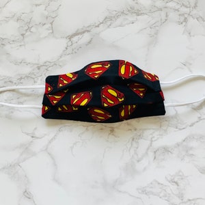 May include: Black face mask with a white elastic band. The mask is decorated with a repeated pattern of red and yellow Superman logos.