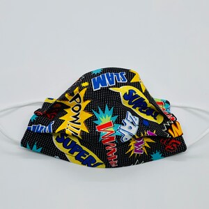 May include: A black face mask with a comic book design. The mask has white elastic ear loops and is covered in colourful speech bubbles with words like "POW!" and "SUPER!" against a black background with white dots.