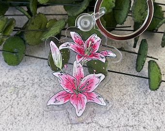 Op de afbeelding: Een transparante acryl sleutelhanger met een handgetekend ontwerp van twee roze lelies met groene stelen en bladeren. De sleutelhanger heeft een zilveren metalen ring.